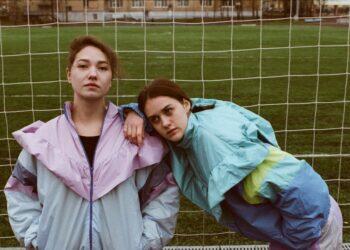 Two women standing in front of football field wearing 90s fashion jackets (Unsplash | Armen Aydinyan)