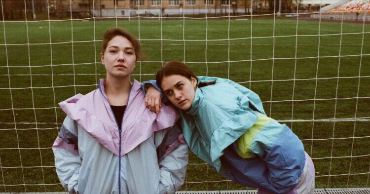 Two women standing in front of football field wearing 90s fashion jackets (Unsplash | Armen Aydinyan)