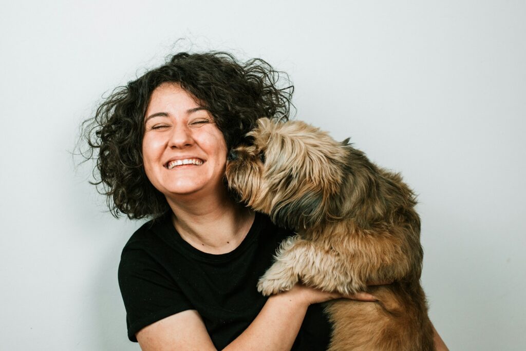 A dog kissing a woman with black curly hair who is wearing a black shirt.
