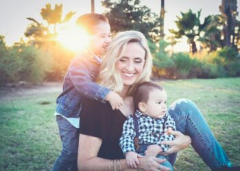 woman holding baby sitting on green grass field under sunset