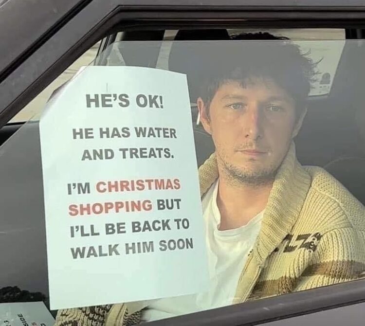 a man in a car with a sign taped to the window that says “he’s ok! he has water and has treats. i’m christmas shopping but i’ll be back to walk him soon.”