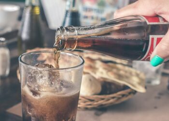 person pouring Coca-Cola soda on clear drinking glass near round white ceramic plate