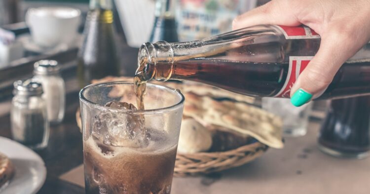 person pouring Coca-Cola soda on clear drinking glass near round white ceramic plate