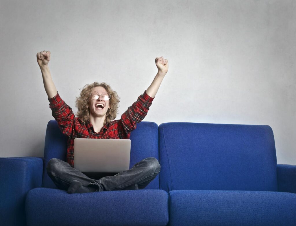 Photo of Excited Person With Hands Up Sitting on A Blue Sofa While Using a Laptop