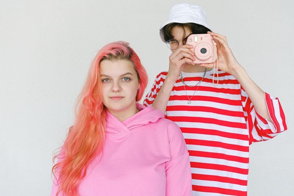 Calm informal teenager couple in bright colorful clothes standing against white background and taking picture on modern instant photo camera