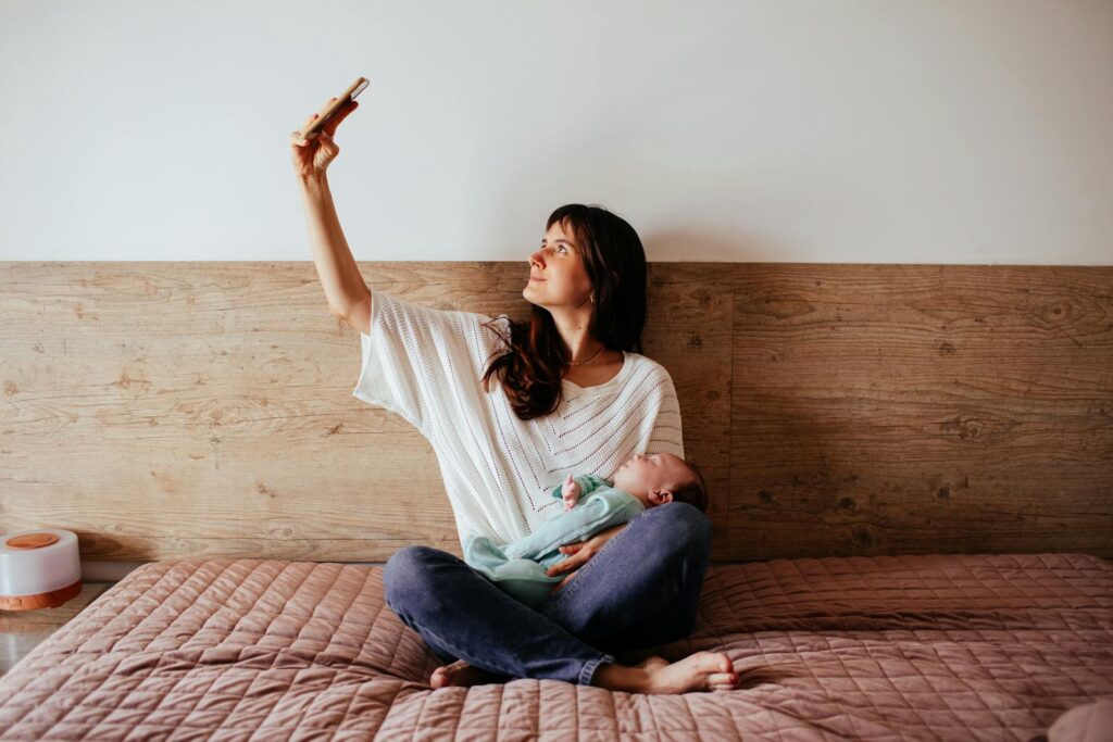 Woman Sitting Cross-Legged and Holding a Baby While Taking a Selfie on a Bed