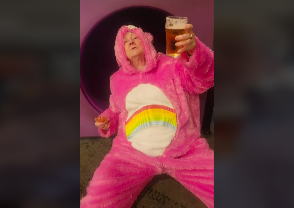 An older woman wearing a care bear onesie holds a cup of beer at the bottom of a slide