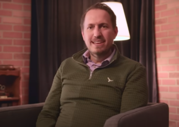 Travis Shreeve in a green sweater sits in front of a background during an interview