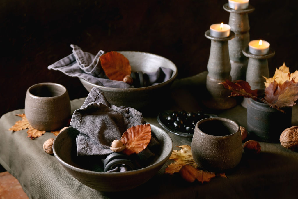 Rustic Autumn Halloween or Thanksgiving table setting with empty craft ceramic tableware, grey bowls and cups, burning candles on linen tablecloth with autumn yellow leaves and acorns, Dark background