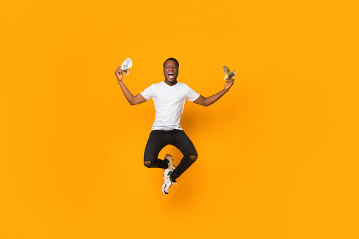 Excited Afro Man Holding Money Jumping In Air, Orange Background