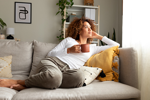 Pensive multiracial woman relaxing at home, sitting on the sofa drinking morning coffee.