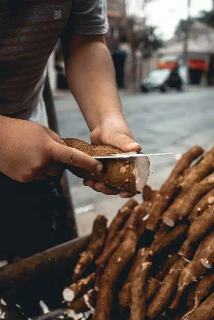 A person peeling cassava with a knife at an outdoor market, showcasing traditional food preparation.