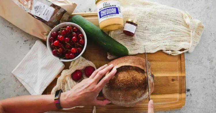 Person cutting bread on cutting board with groceries scattered on the table (Unsplash | Grace O'Driscoll)